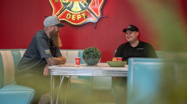 Två bilservice-män sitter vid ett bord och fikar, rummet har 50-talsinredning.